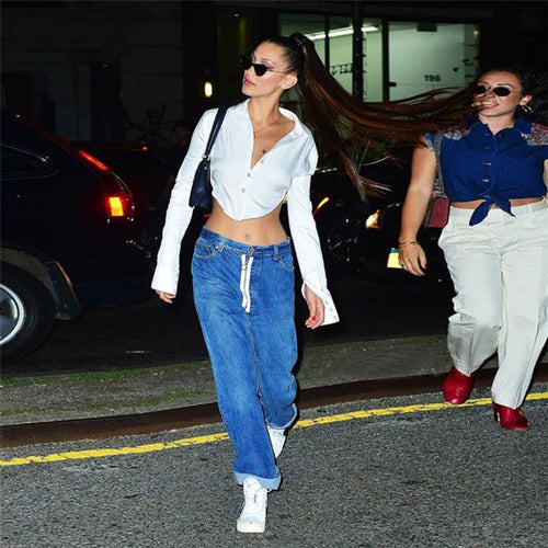 Woman with long dark hair in a high ponytail wearing black sunglasses, walking on a city street at night. She models a white long-sleeve cropped button-up shirt tied at the back, exposing her midriff, paired with blue cuffed wide-leg jeans and white sneakers while carrying a black shoulder bag.