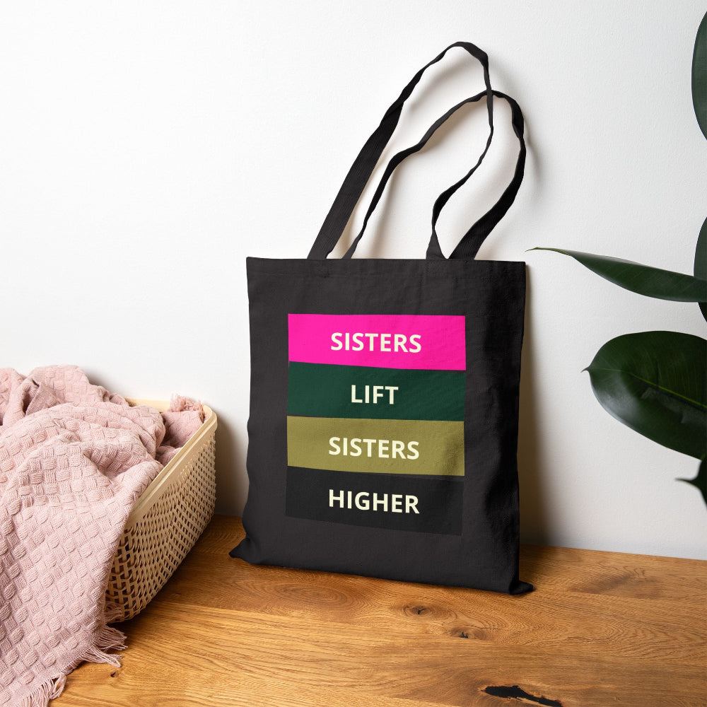 Black canvas tote bag with "SISTERS LIFT SISTERS HIGHER" graphic on wooden table with pink throw and plant.