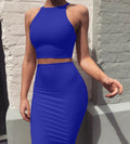 Woman in vibrant blue halter crop top and high-waisted midi skirt set posing against white brick wall.
