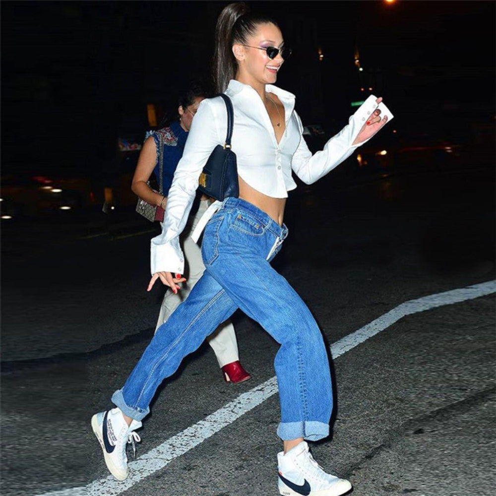 Woman with long dark hair in a high ponytail wearing black sunglasses, smiling while walking on a city street at night. She is wearing a white long-sleeve cropped button-up shirt tied at the back for an open-back look, blue wide-leg cuffed jeans, and white high-top sneakers with black Nike swoosh.