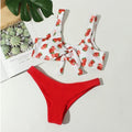 Strawberry-patterned bikini top and red bottom on a white surface with a plant leaf and paper in the background.