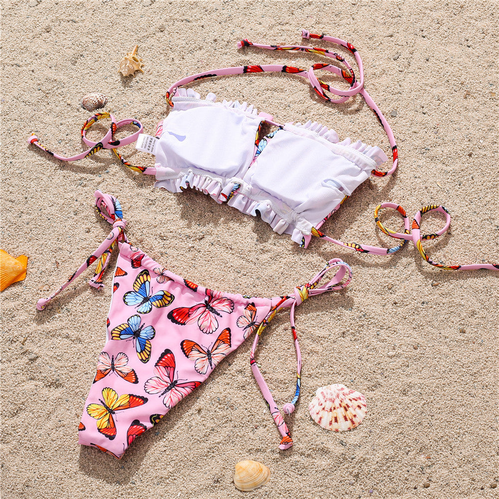 White and pink butterfly print bikini set on sand – trendy ruffled top with colorful tie bottoms, beach flat lay swimwear.