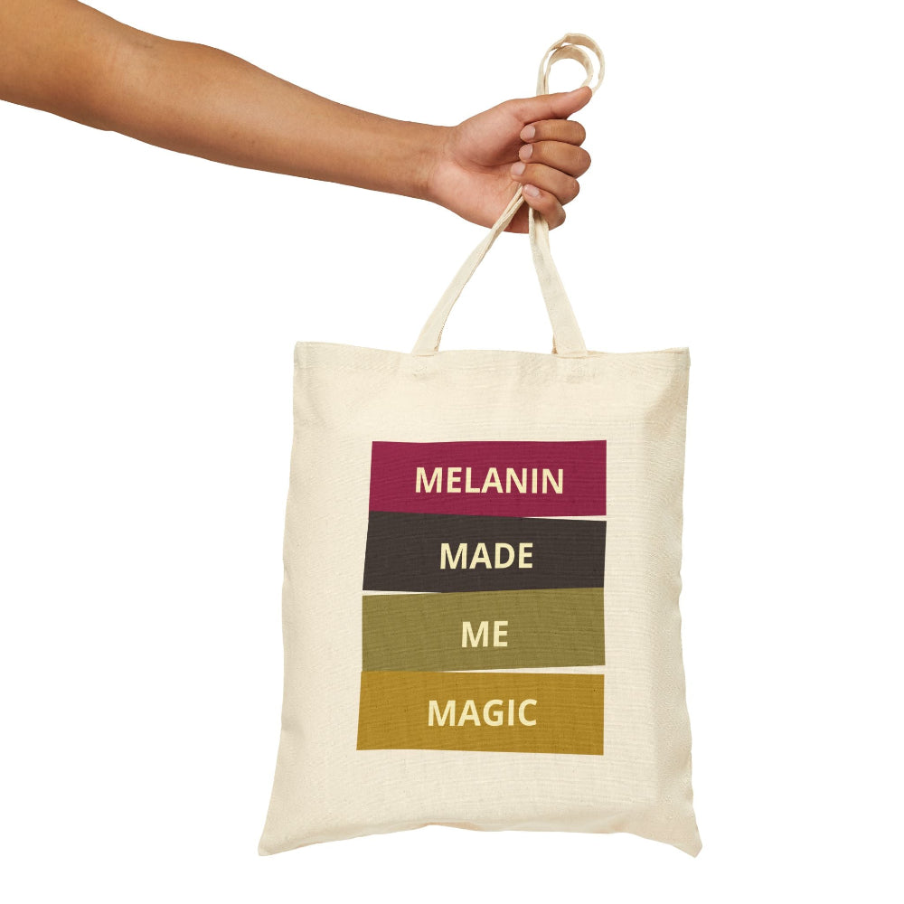Hand holding natural canvas tote bag with "MELANIN MADE ME MAGIC" colorful block graphic.