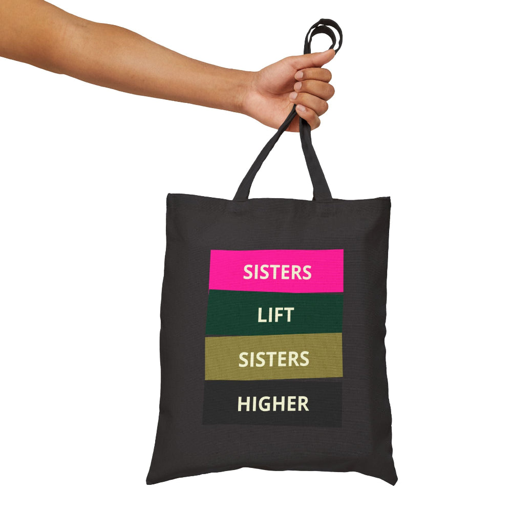 Hand holding black canvas tote bag with "SISTERS LIFT SISTERS HIGHER" graphic in colorful blocks.