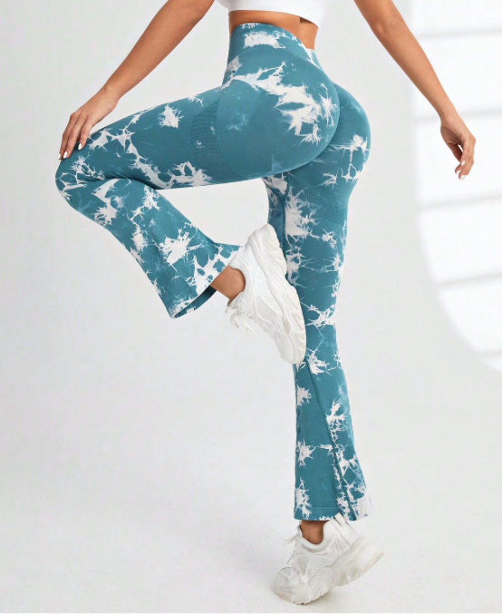 Woman modeling teal and white tie-dye high-waisted flared pants, shown from a rear three-quarter view with one leg lifted, wearing white sneakers against a clean white studio background.