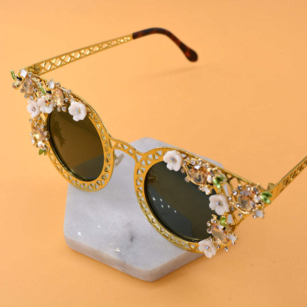 Elegant gold metal cat-eye sunglasses featuring intricate lattice frames, white floral accents, green leaves, sparkling crystals, and dark green-brown tinted lenses, displayed on marble