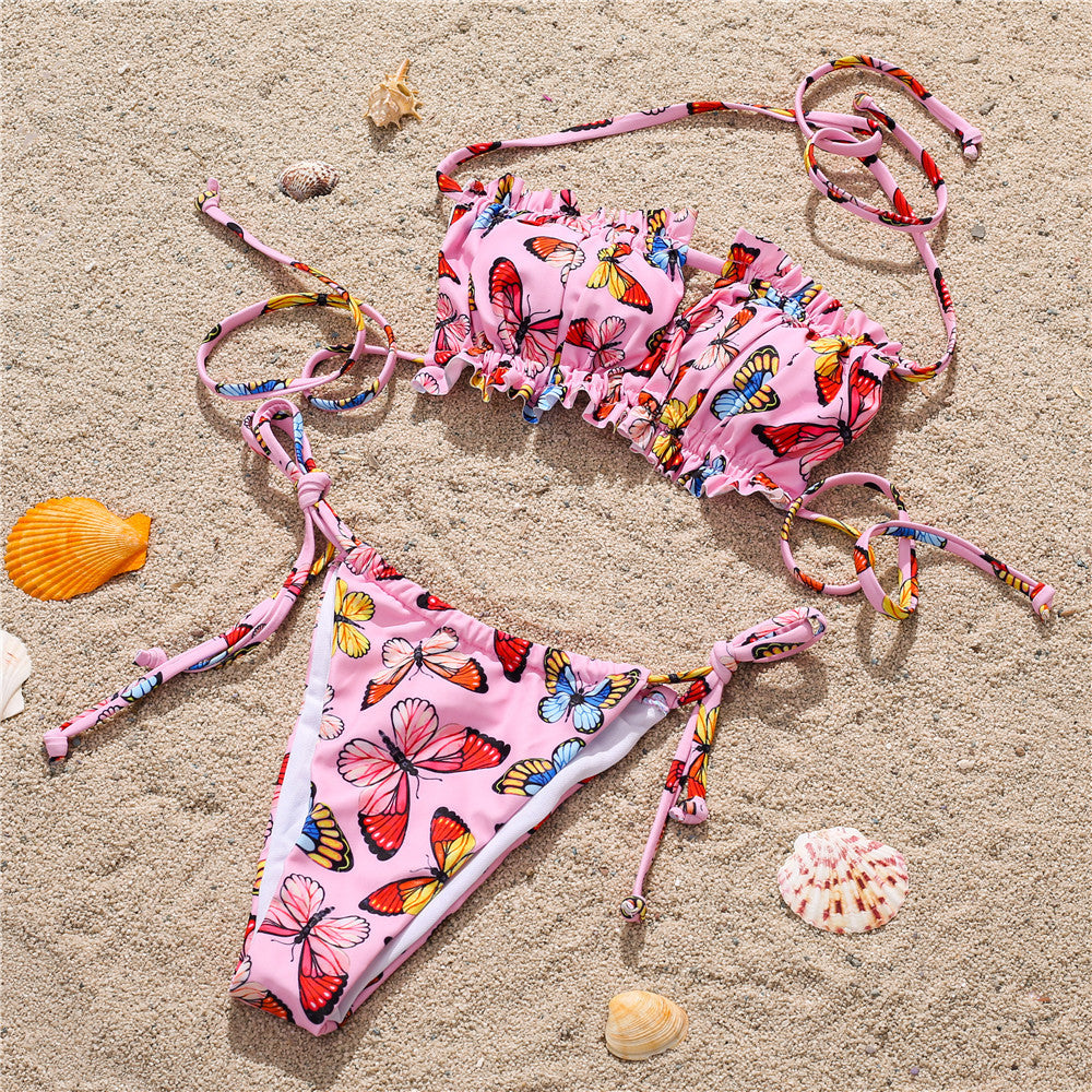 Pink butterfly print bikini on sand – trendy colorful ruffled swimwear set with ties, beach product flat lay.
