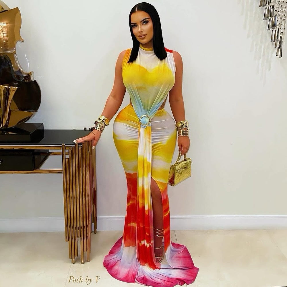 "Woman with long black hair wearing a vibrant yellow, orange, and pink tie-dye sleeveless maxi dress with high neck, front cut-out detail, and side slit, posing with hand on a gold-accented table against a white wall."