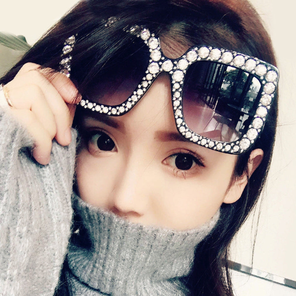 Model holding black oversized square sunglasses with crystal-studded frames and dark lenses on her forehead.
