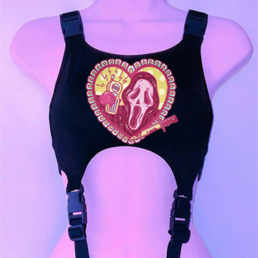 Black harness-style crop top on a mannequin with adjustable buckle straps and large side cut-outs. Features a bold heart-shaped graphic with Ghostface from Scream, a phone with lightning bolts, a bloody knife, and horror-themed details on the chest, displayed under purple lighting.