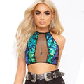 Woman with long wavy blonde hair modeling a vibrant iridescent sequin halter crop top featuring blue, green, and purple sequins with a sheer black fishnet mesh panel at the center. Styled with a wide black statement belt with large ornate silver buckles and matching dangling earrings.