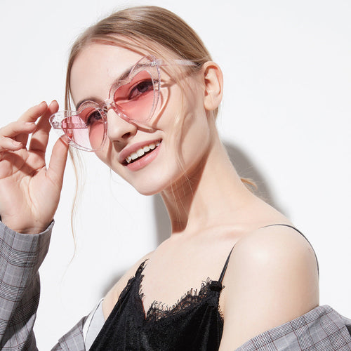 Smiling blonde woman wearing pink glittery heart-shaped sunglasses with translucent frames, adjusting them with one hand while dressed in a black lace-trimmed top and plaid blazer against a bright white background