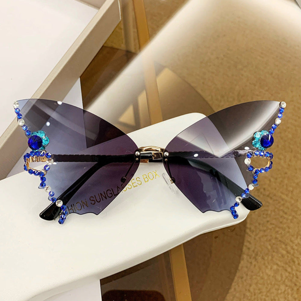 Rimless butterfly sunglasses with dark gray lenses and blue crystal wing details, displayed on a white glasses case.