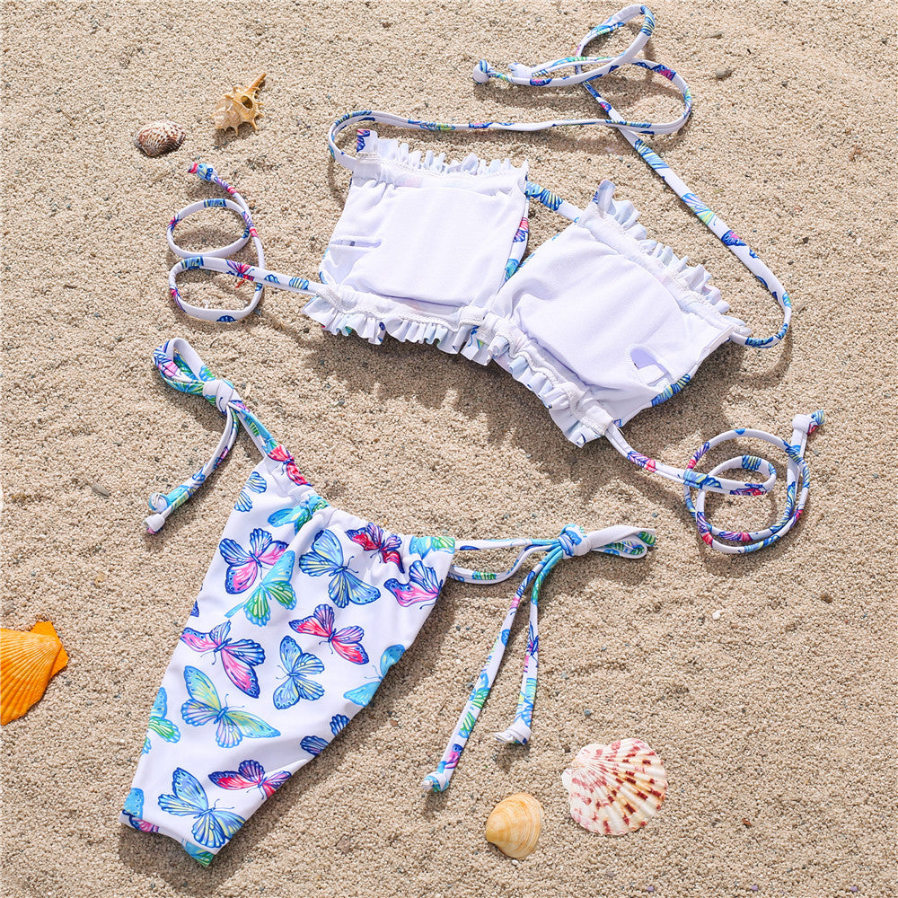 White butterfly print bikini set on sand – trendy ruffled swimwear with colorful ties, beach product flat lay.
