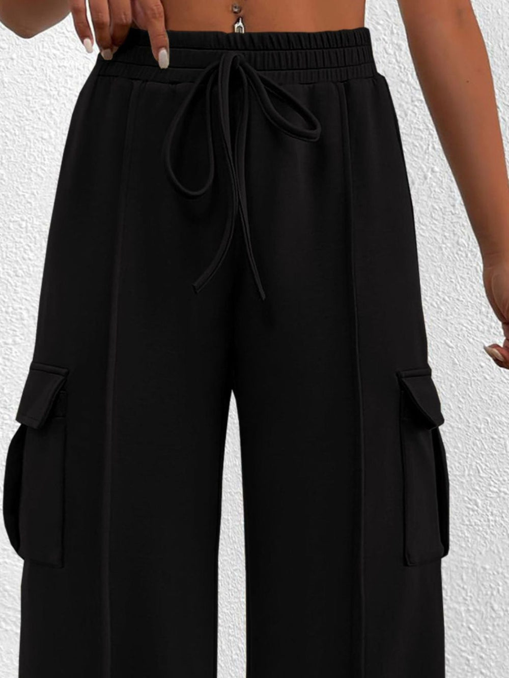 Woman wearing black wide leg cargo pants with drawstring waist and large side pockets against a textured white background