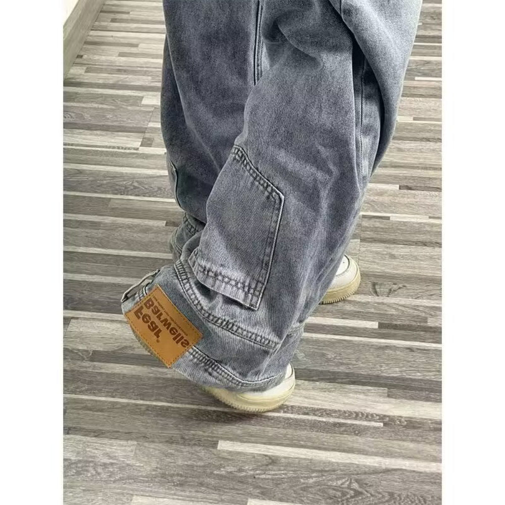 women wearing relaxed-fit blue denim cargo-style jeans featuring a prominent leather patch/label at the cuff/hem area. Detail shot from a high angle showing the fabric texture, stitching, and fit against light wood flooring.