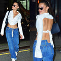 Two-panel collage of a woman with long dark hair in a high ponytail wearing black sunglasses. She models a white long-sleeve cropped button-up shirt tied at the back for an open-back cut-out effect, exposing her midriff and back. Styled with blue wide-leg jeans in both front and back views.