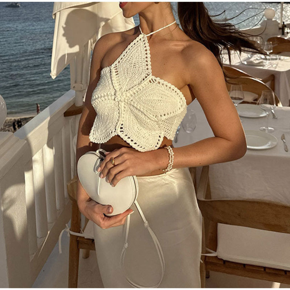 Model wearing a white crochet halter crop top with intricate floral butterfly design, paired with a white satin skirt and holding a white heart-shaped bag on a luxury seaside terrace.
