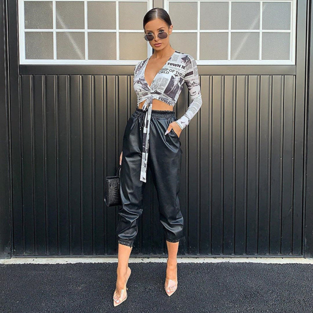 Black and white newspaper print long sleeve tie-front crop top with deep plunge V-neck, paired with black faux leather jogger pants and clear heels, worn by a model.