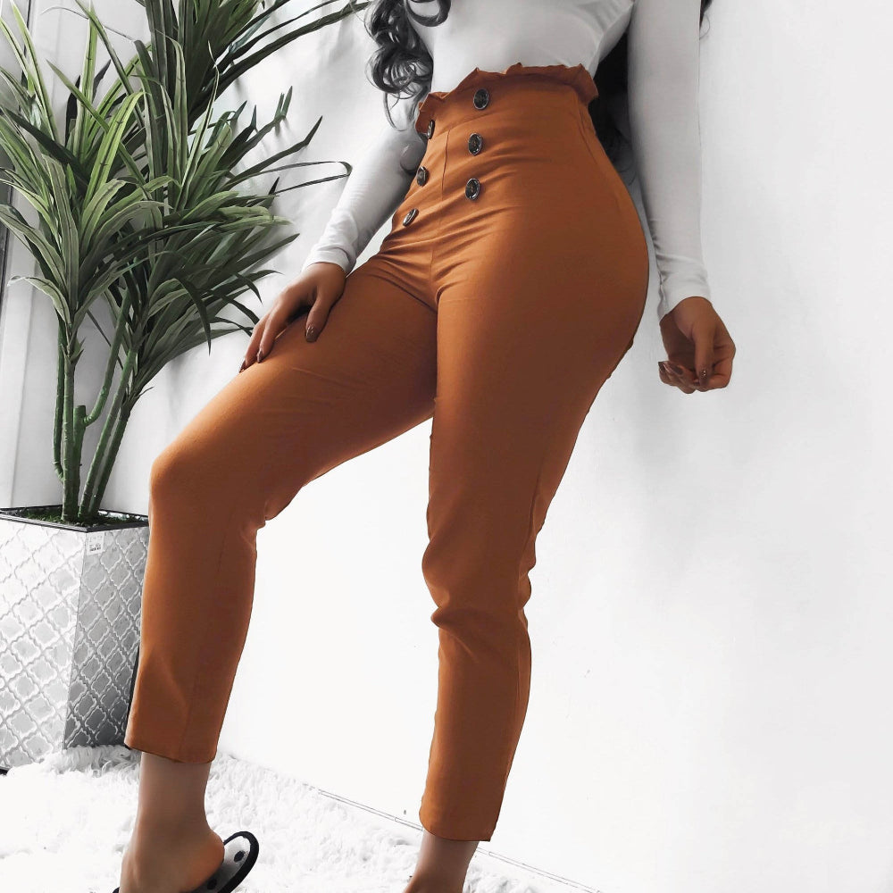 Woman wearing orange high-waisted pants with decorative front buttons, paired with a white long-sleeve top, posing indoors next to a potted plant.