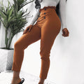 Woman wearing orange high-waisted pants with decorative front buttons, paired with a white long-sleeve top, posing indoors next to a potted plant.