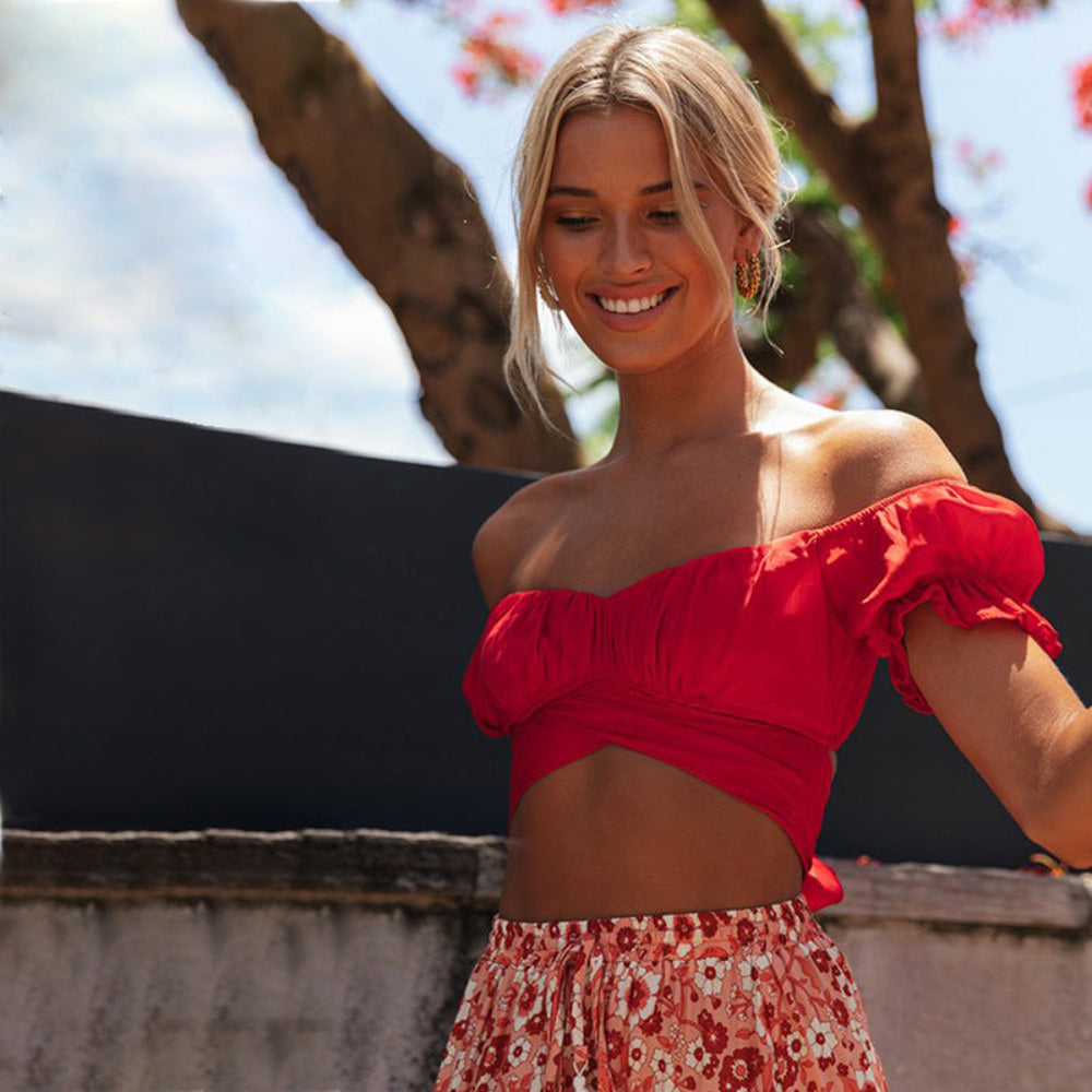 Red off-shoulder ruched crop top with puff sleeves and front cut-out detail, paired with a floral skirt, worn by a smiling blonde model outdoors.