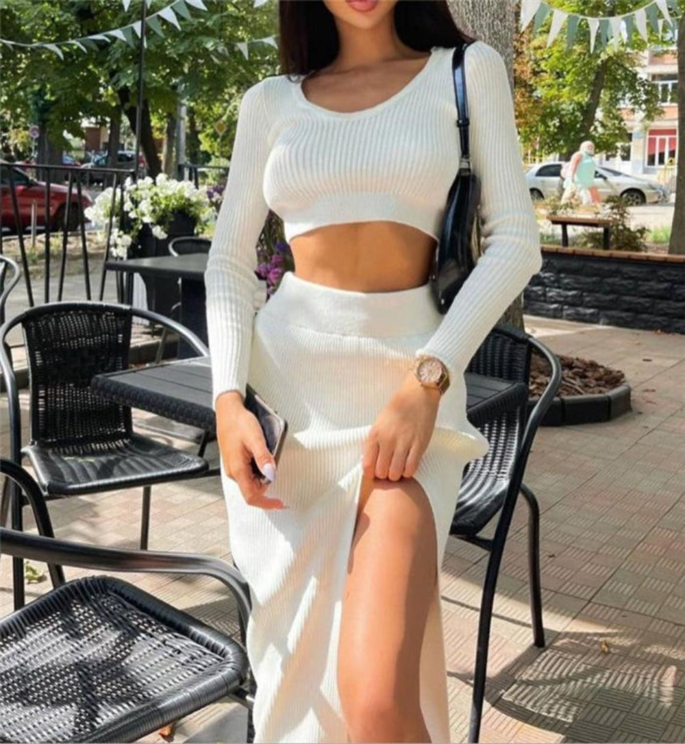 Woman wearing white ribbed long-sleeve crop top and high-slit midi skirt set, standing outdoors at cafe setting.