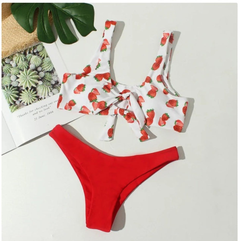 Strawberry-patterned bikini top and red bottoms on a white surface with a plant leaf and paper in the background.