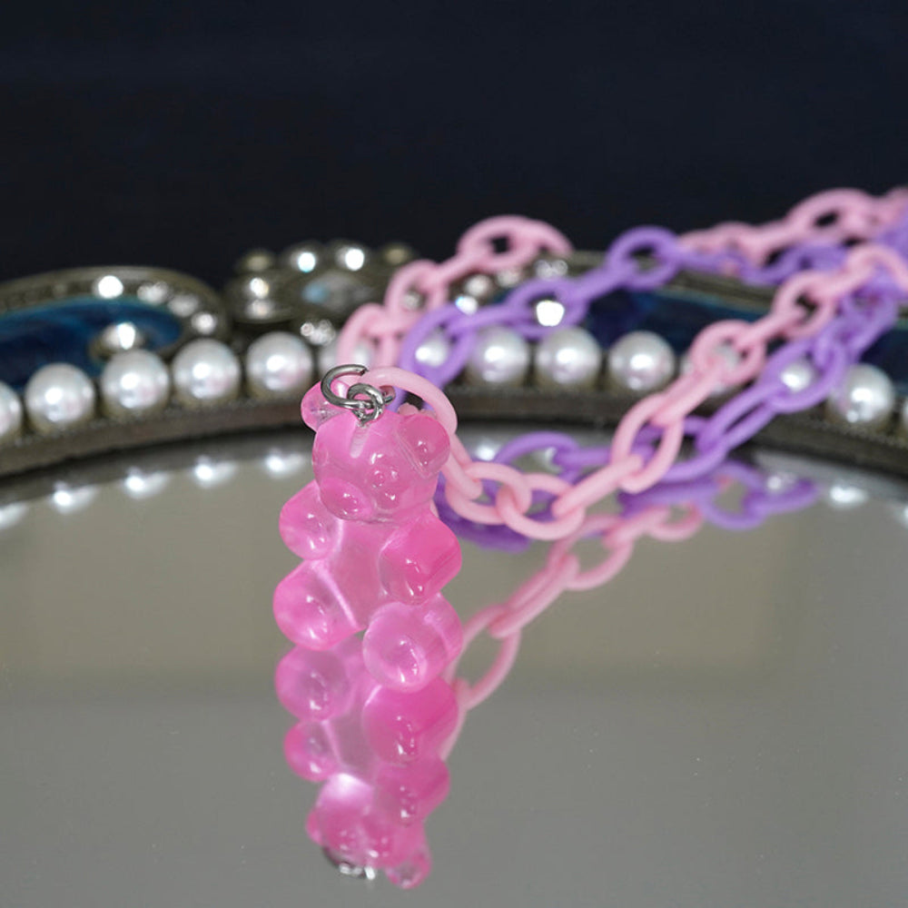Close-up of a translucent hot pink gummy bear pendant attached to a pastel pink and purple chain necklace. The playful charm is shown on a reflective mirror surface with a decorative pearl-accented frame in the background, highlighting its glossy, fun details.