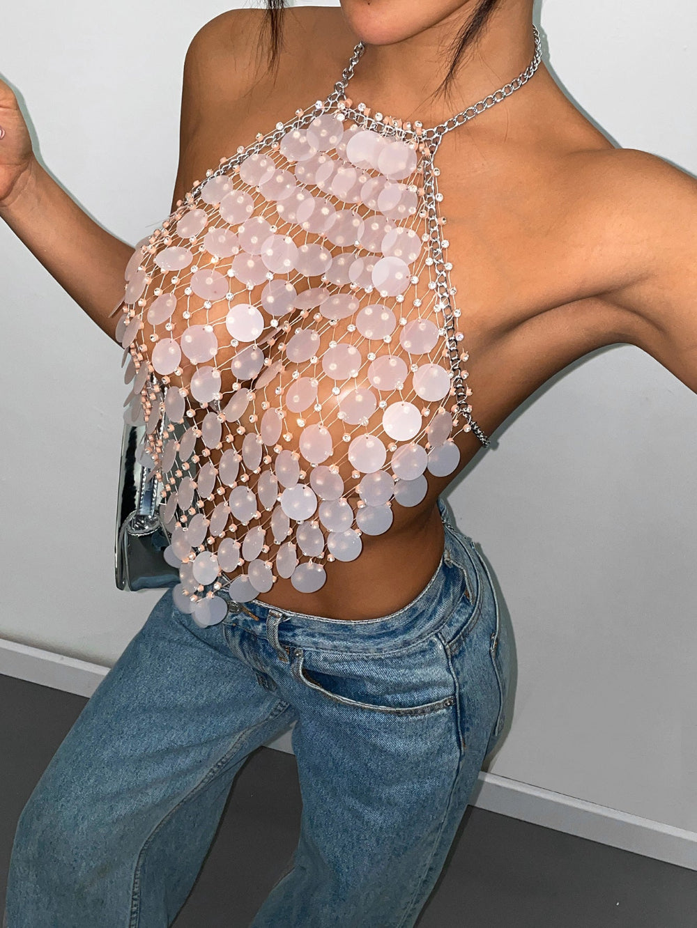 Woman wearing a stunning pale pink sequin halter crop top with cascading layers of iridescent circular sequins and silver chain straps. Styled with blue denim jeans in a dynamic pose with arms raised.