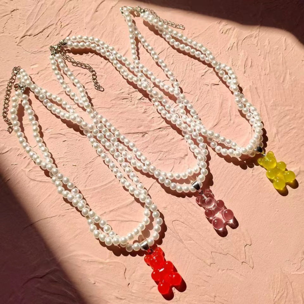 Three delicate multi-strand pearl necklaces featuring colorful gummy bear pendants in vibrant red, translucent pink, and bright yellow. The necklaces are elegantly displayed on a textured pink background with natural sunlight casting soft shadows.