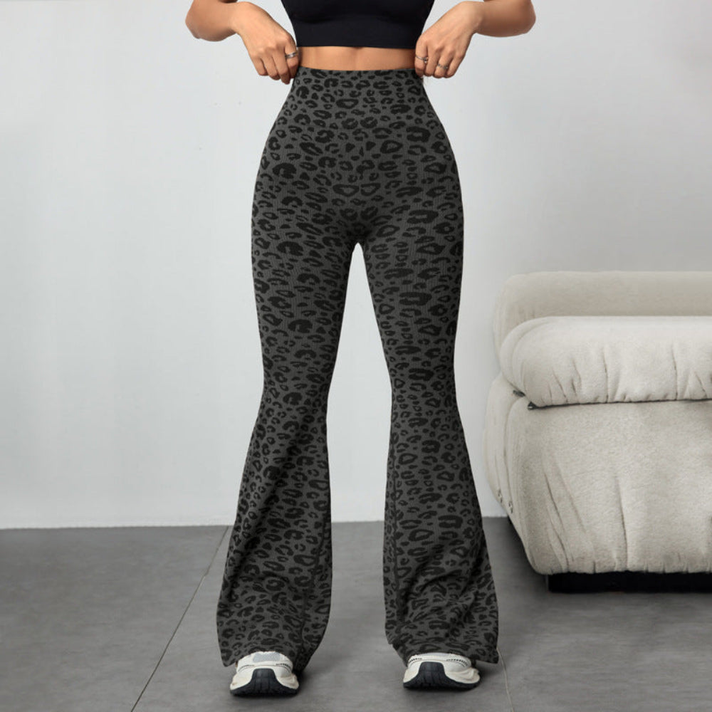 Woman modeling black leopard print high-waisted flared pants, standing and adjusting the waistband. Paired with a black crop top and sneakers in a bright indoor setting with a gray couch.