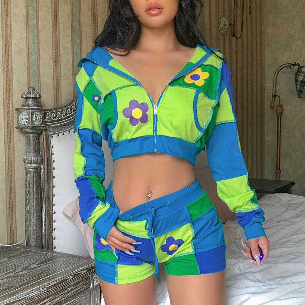 Woman modeling a vibrant patchwork cropped zip-up hoodie and matching shorts set in lime green, blue, and green color-block panels. The hoodie features a hood, front zipper, patch pockets, and colorful daisy flower appliqués, while the shorts have a drawstring waist and coordinating flower details. Styled with a visible belly button piercing.