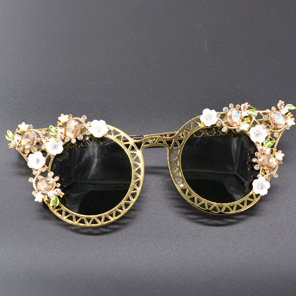 Vintage-style gold metal cat-eye sunglasses featuring intricate lattice frames, delicate white floral accents, green leaves, and sparkling crystal details on black round lenses