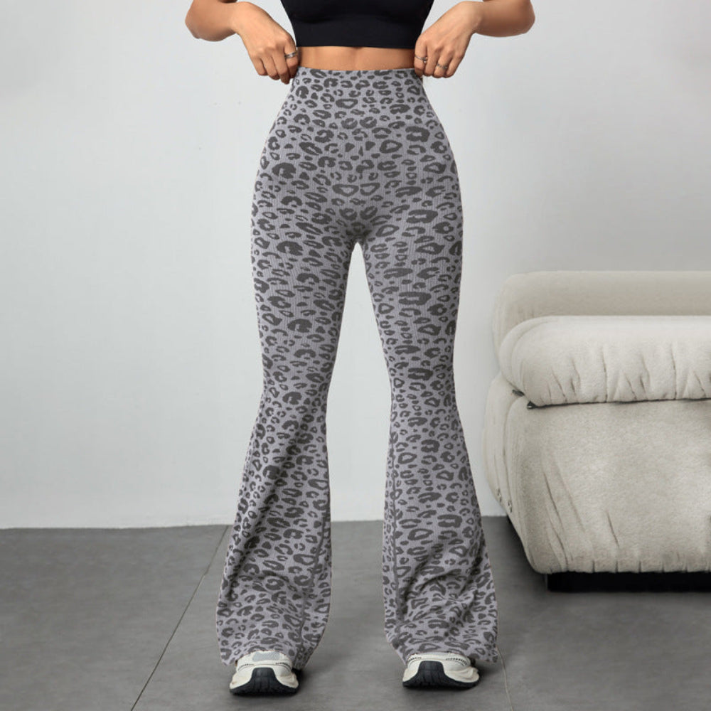 Woman modeling gray leopard print high-waisted flared pants, standing and adjusting the waistband. Paired with a black crop top and sneakers in a bright indoor setting with a gray couch.