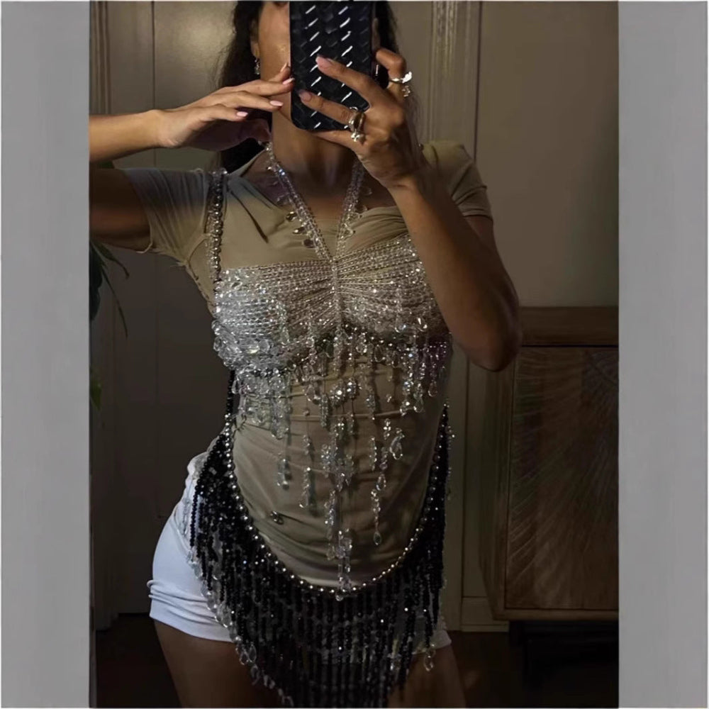 Woman taking a mirror selfie wearing a beige short-sleeve bodysuit under a dramatic silver and black crystal beaded fringe top with heavy cascading bead strands and sparkling embellishments. Styled with white mini shorts.