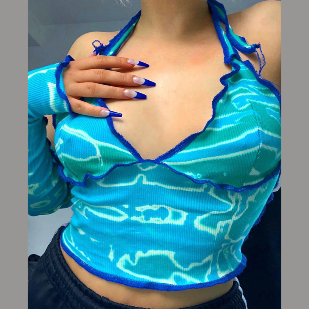 Close-up of a woman wearing a vibrant turquoise and blue ribbed knit halter crop top with white wave pattern, deep plunging neckline, and ruffled scalloped edges trimmed in blue. She has long blue and white French tip nails resting on her chest, paired with a matching long sleeve.