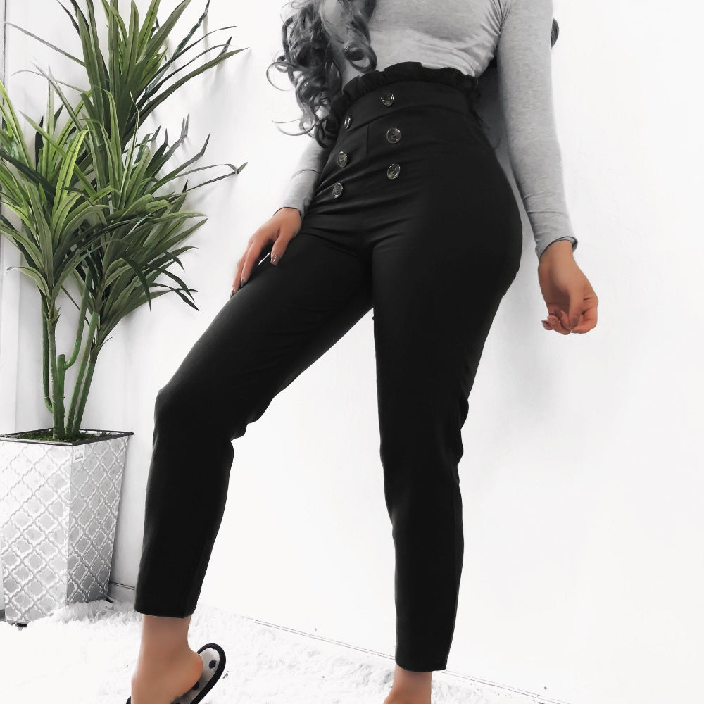 Woman wearing black high-waisted pants with decorative front buttons, paired with a gray long-sleeve top, posing indoors next to a potted plant.