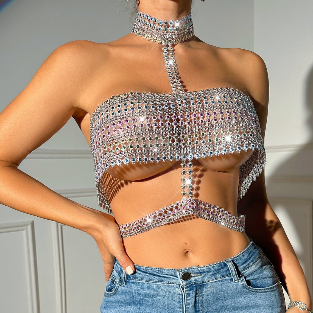 Woman wearing a dazzling iridescent rhinestone halter crop top with high choker neck, multicolored crystal embellishments, and sparkling sequin details. Styled with light blue ripped jeans, hand on hip, showcasing the intricate sparkling design.