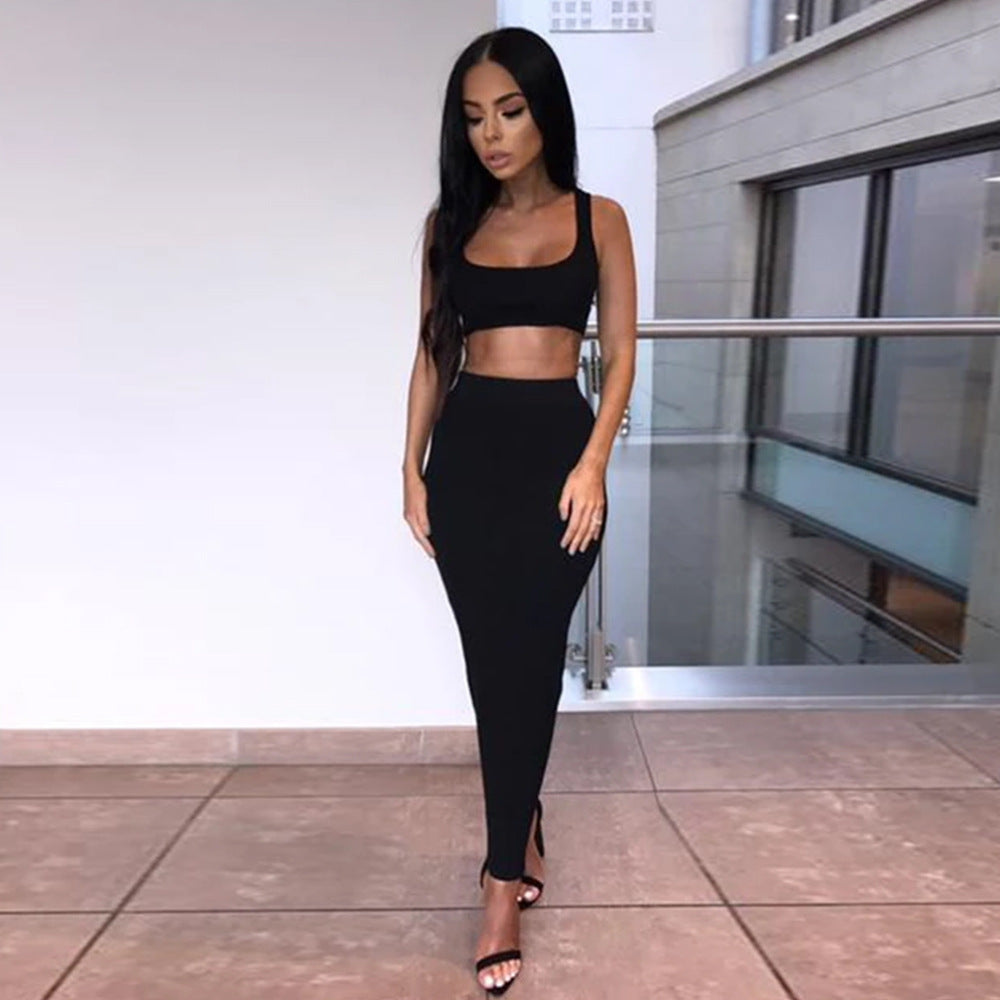 Model in a matching black two-piece outfit featuring a sleeveless crop top and form-fitting high-waisted midi skirt. Full-body fashion pose standing confidently in a bright contemporary interior with white walls, glass railing, and neutral tiled floor.