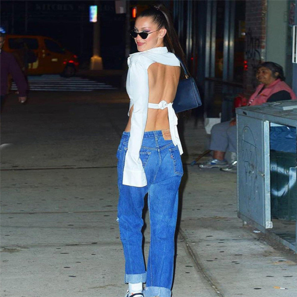 Woman with long dark hair in a high ponytail wearing black sunglasses, modeling a white long-sleeve cropped button-up shirt tied at the back for an open-back cut-out effect that exposes her midriff. Paired with blue cuffed jeans and a dark blue shoulder bag on a city street at night.