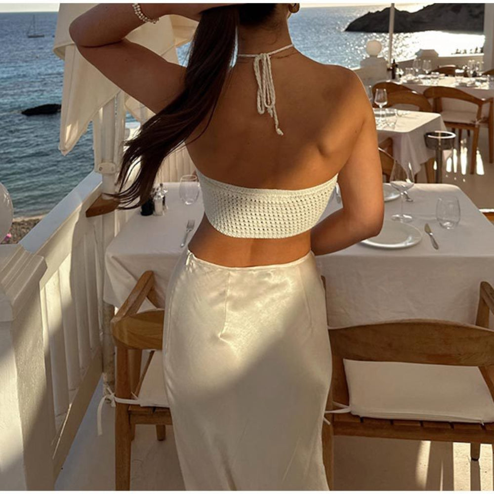 Back view of model wearing a white crochet halter crop top with open back and tie closure, paired with a white satin skirt, standing on a luxury seaside terrace.