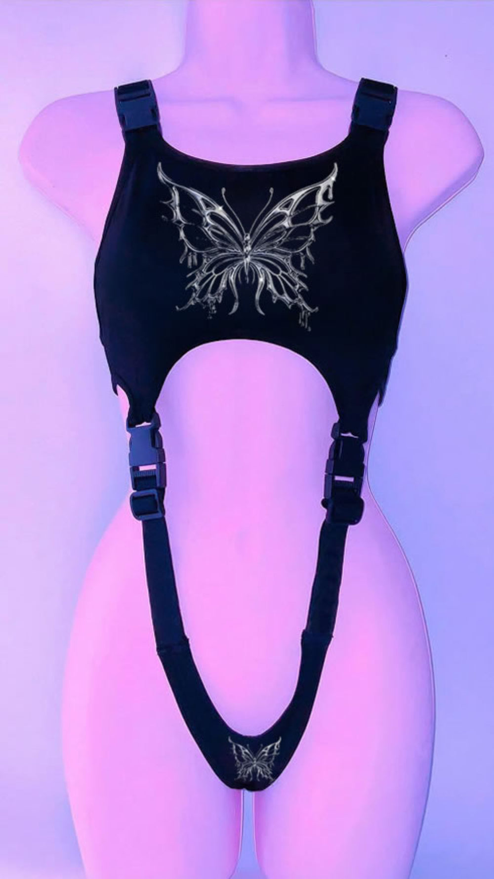Black harness-style crop top on a mannequin with adjustable buckle straps and side cut-outs. Features a large ornate silver butterfly graphic on the chest and a smaller matching butterfly at the bottom, displayed under purple lighting.