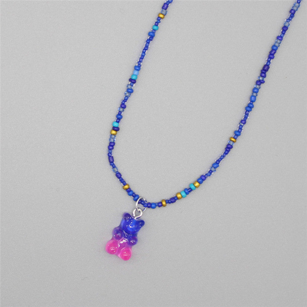 Blue beaded necklace featuring various shades of blue seed beads with gold accents and a sparkling translucent gummy bear pendant in a purple-to-pink gradient. Playful single-strand design laid flat on a light gray background, showcasing its vibrant and whimsical style.