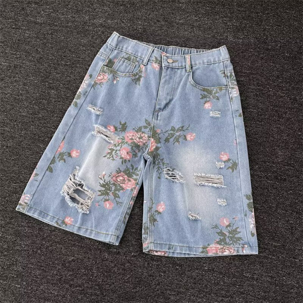 Light blue distressed denim shorts with pink floral rose prints, distressed rips, and frayed details, laid flat on a dark surface. Vintage-inspired fashion product photography for Boutique Jolie.