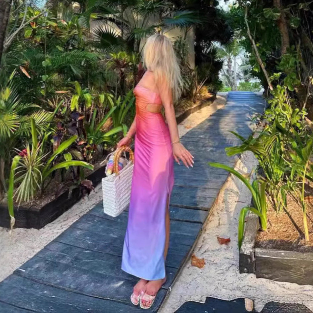 Model in a beautiful ombre strapless floor-length dress transitioning from bright pink at the bust to soft purple at the hem. Side/rear view as she walks barefoot on a wooden path through dense tropical foliage, carrying a white woven tote bag.