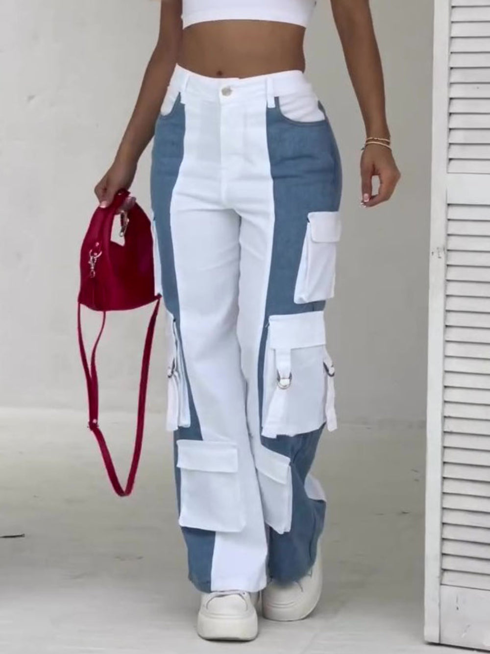 Front view of a woman wearing high-waisted color-block cargo jeans in white and blue denim panels, featuring multiple large cargo pockets with flaps and metal hardware. Styled with a white crop top, white sneakers, and holding a small red crossbody bag.