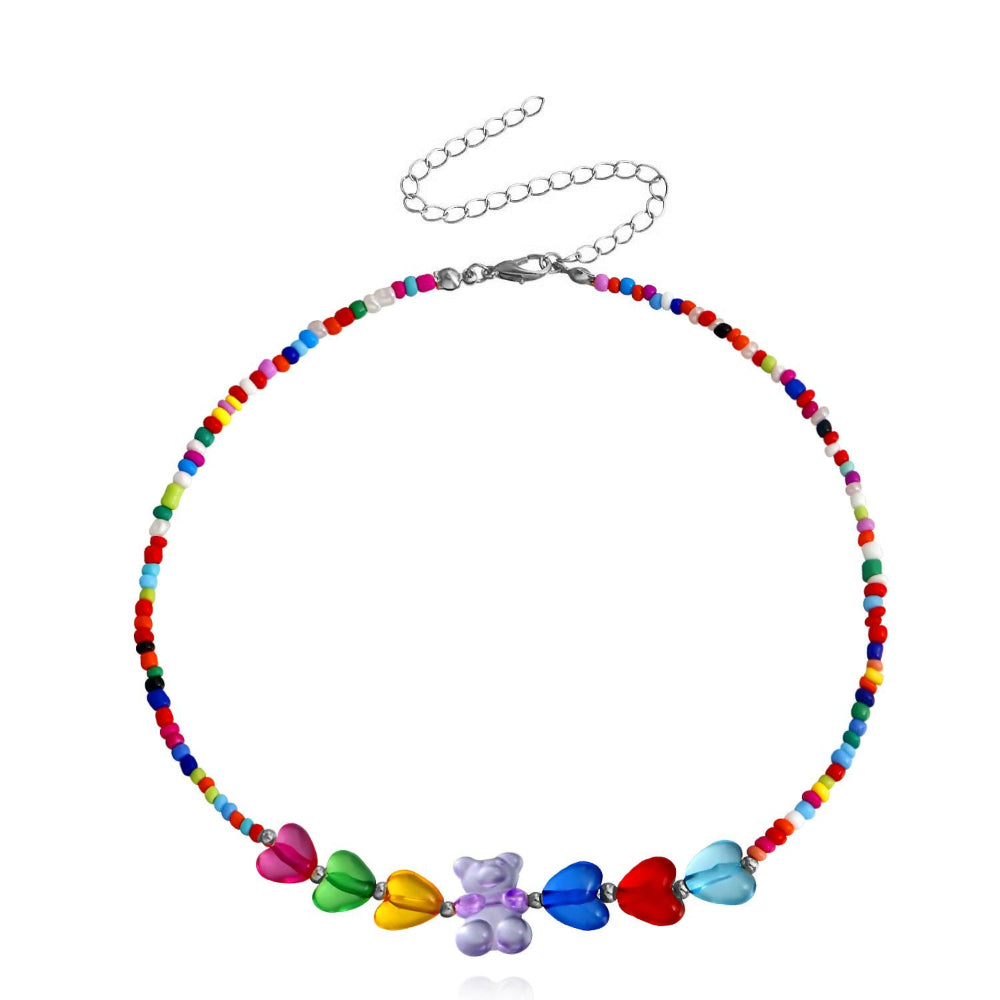 Rainbow beaded choker necklace with multicolored seed beads and large translucent heart-shaped beads in pink, green, yellow, blue, red, and light blue. Features a central purple gummy bear charm and silver lobster clasp with extender chain. Fun, vibrant statement piece displayed on a clean white background.