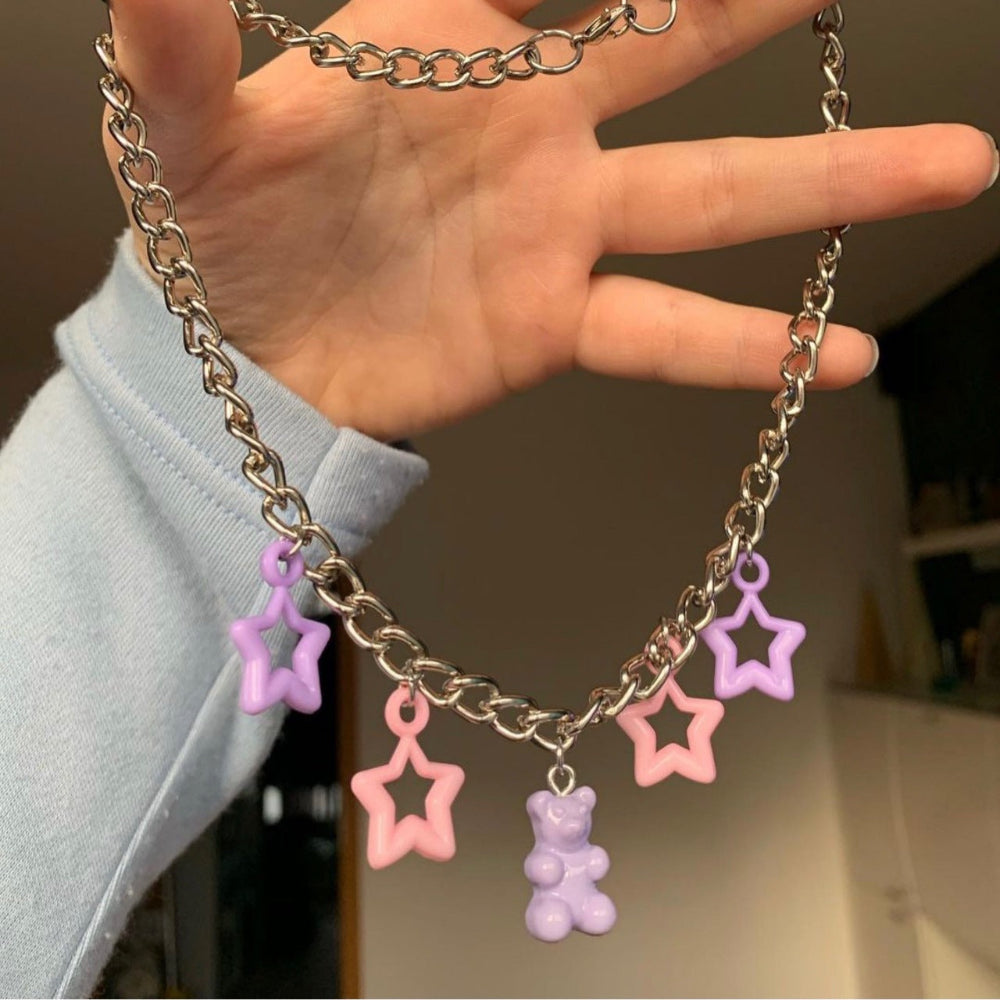 Silver curb chain necklace with playful pastel charms — purple and pink star pendants plus a purple gummy bear charm. Held in hand to show the chunky chain and dangling details, creating a fun Y2K-inspired statement piece.