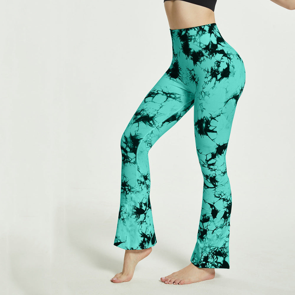 Woman modeling teal and black tie-dye high-waisted flared pants, shown from a side view while standing barefoot against a clean white studio background.